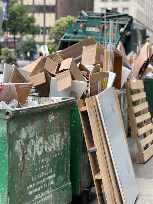 A large green metal rubbish skip filled with a mixture of debris, including numerous cardboard boxes, some flattened and others still in their original shape, along with a few foam packaging pieces. T